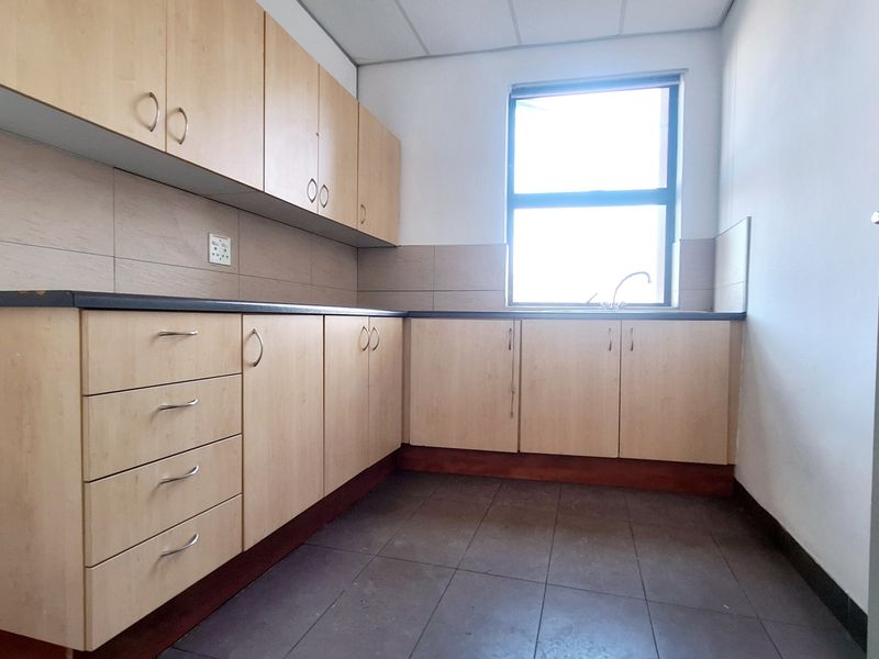 A functional office kitchenette featuring light wood-finish cabinetry, dark laminate countertops, a stainless steel sink, and tiled flooring.