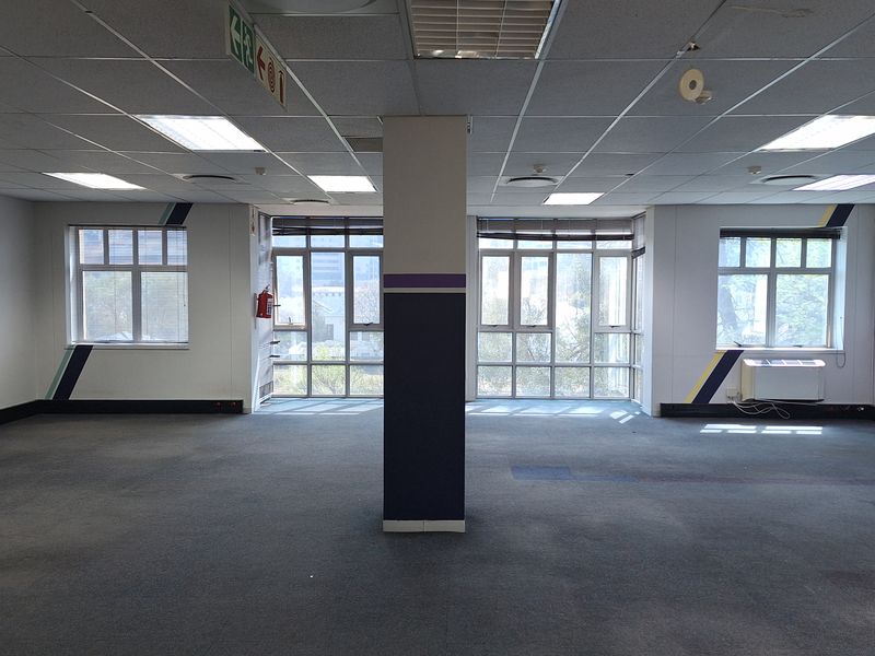 An open-plan office space featuring a central structural column, suspended ceiling with recessed lighting, and large windows providing natural light.