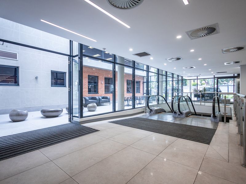 Modern office building entrance lobby featuring floor-to-ceiling glass walls, tiled flooring, and internal escalators leading to other levels.