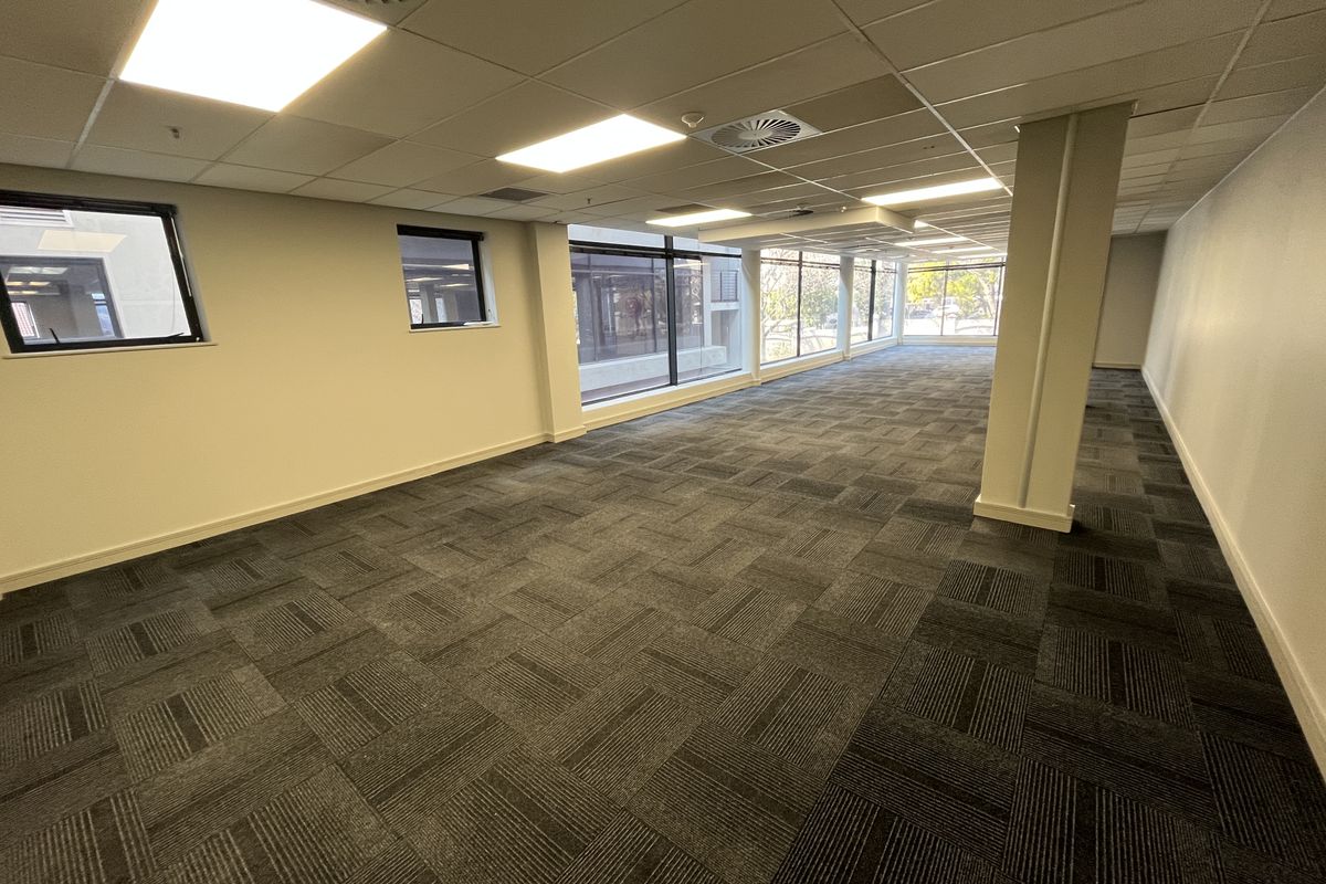 A spacious, open-plan office area featuring neutral walls, carpeted flooring, recessed ceiling lighting, and large windows providing ample natural light.