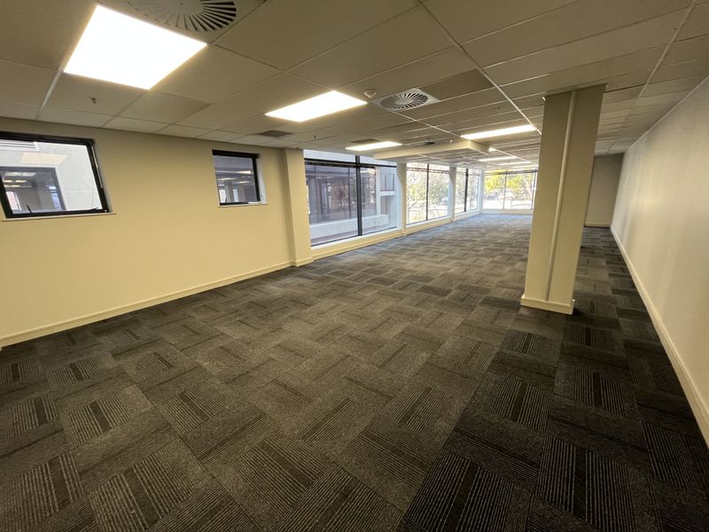 A spacious, open-plan office area featuring neutral walls, carpeted flooring, recessed ceiling lighting, and large windows providing ample natural light.