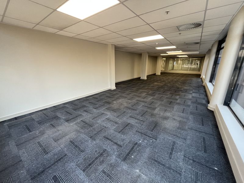 A spacious, empty open-plan office area featuring modern carpet tiles, a suspended ceiling with recessed lighting, and large windows providing natural light.