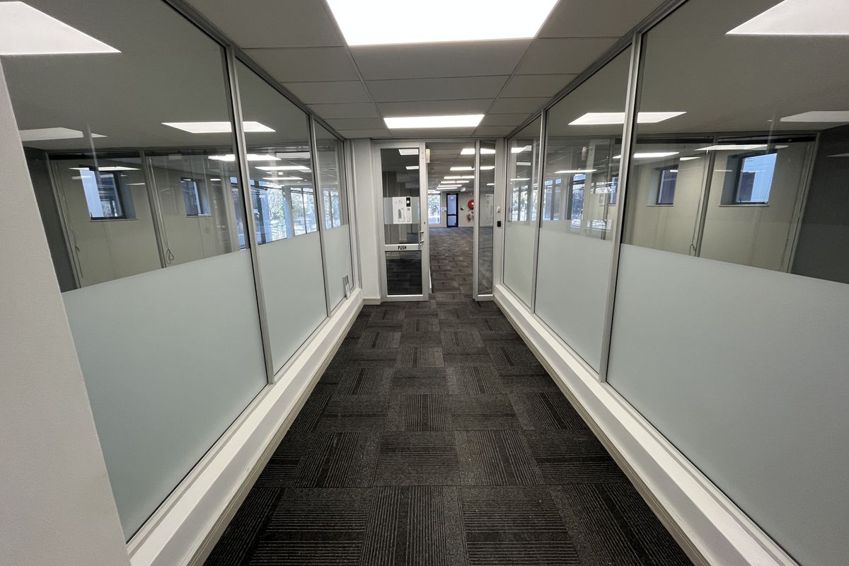 A modern office hallway featuring glass partition walls, carpet tile flooring, and recessed ceiling lighting, providing a clean and professional workspace transition.