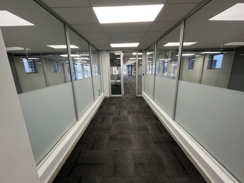 A modern office hallway featuring glass partition walls, carpet tile flooring, and recessed ceiling lighting, providing a clean and professional workspace transition.