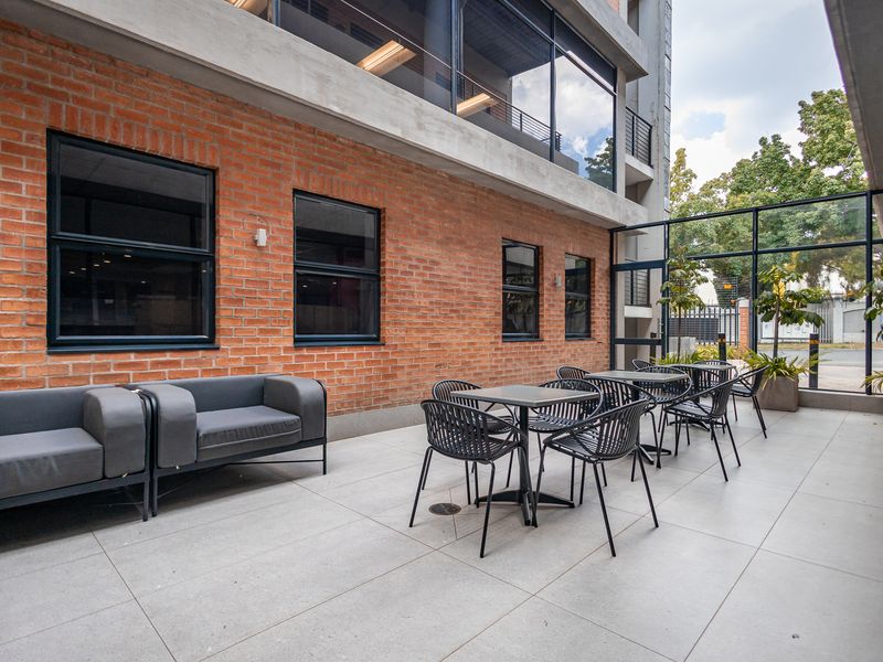 A modern outdoor patio area featuring exposed brickwork, large glass windows, and tiled flooring, providing a professional and inviting space for office occupants.