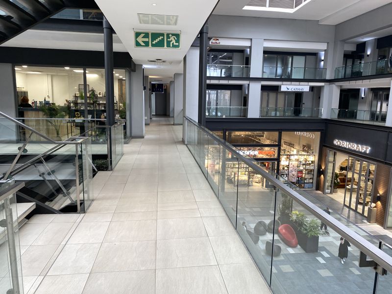 Modern office building atrium featuring a tiled walkway, glass balustrades, and a central staircase overlooking the lower retail level.