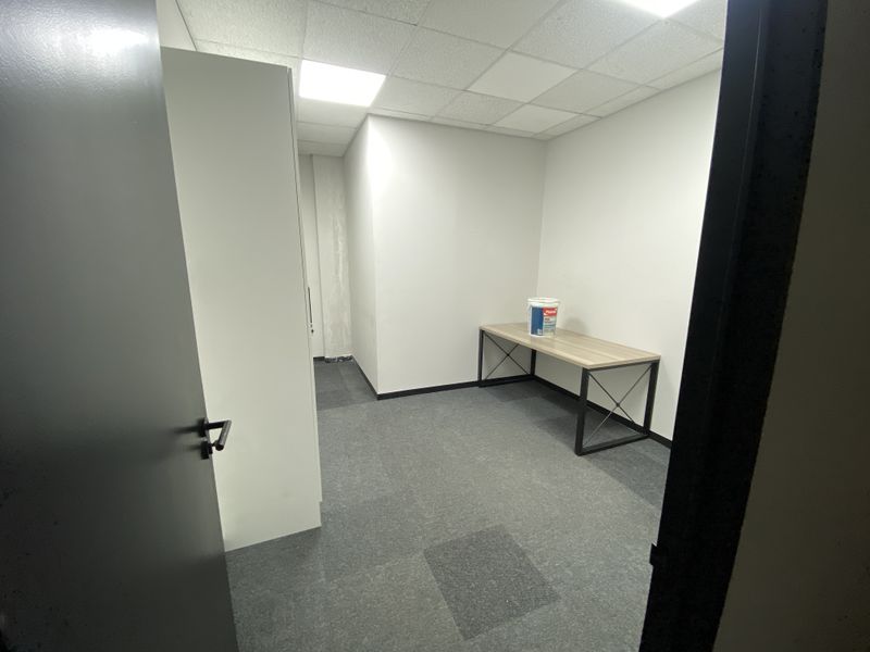 A compact, empty office space featuring carpet tiles, a suspended ceiling with recessed lighting, and neutral wall finishes.