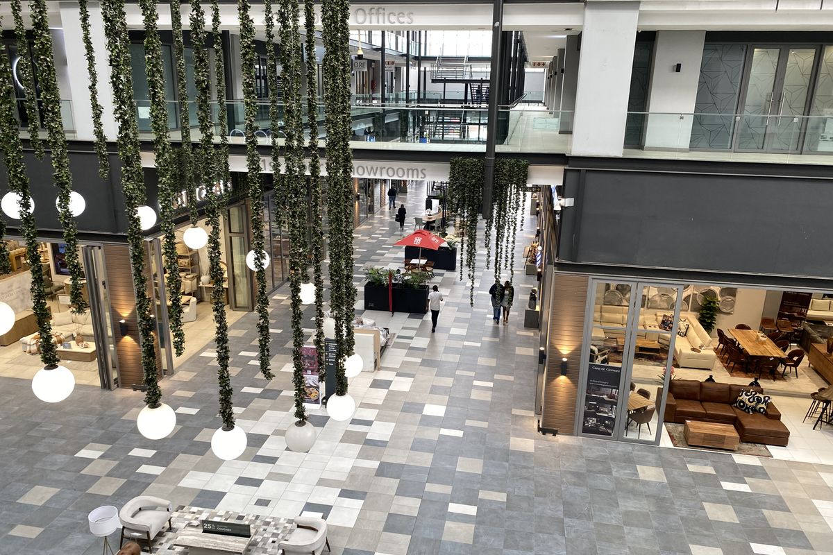 A modern, multi-level commercial atrium featuring tiled flooring, glass-fronted office and showroom spaces, and decorative suspended lighting.