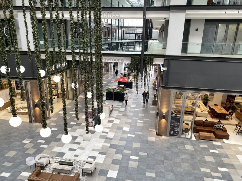 A modern, multi-level commercial atrium featuring tiled flooring, glass-fronted office and showroom spaces, and decorative suspended lighting.