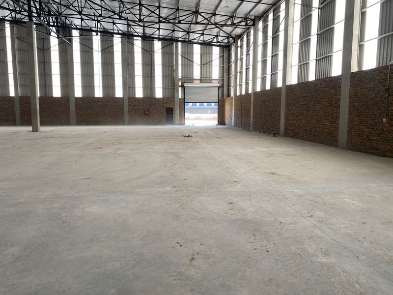 Spacious industrial warehouse featuring high ceilings with exposed steel trusses, concrete flooring, face brick walls, and a large roller shutter door.