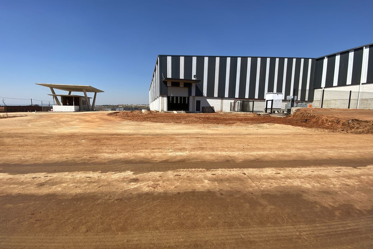 Industrial warehouse facility featuring metal cladding, a roller shutter door, and a separate security gatehouse, set within an expansive, unfinished site.