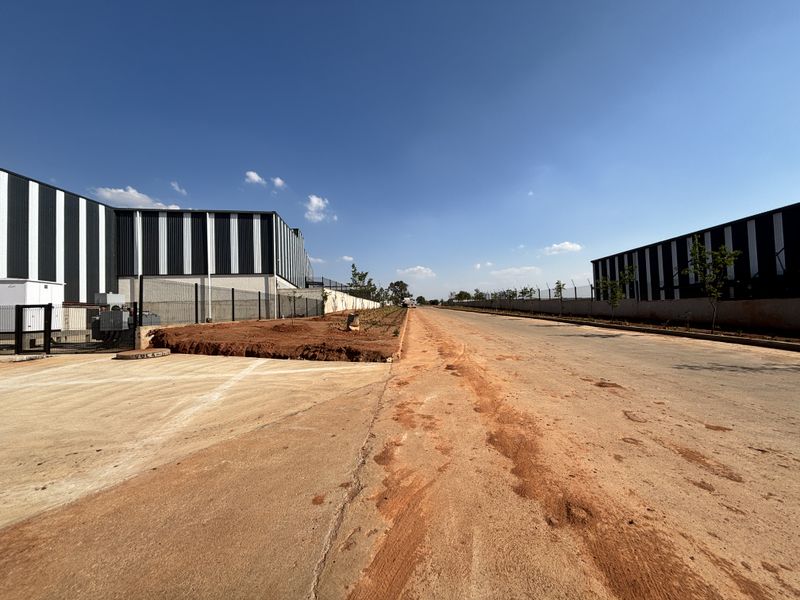 View of an industrial park access road flanked by modern warehouse buildings with perimeter fencing and ongoing site development.