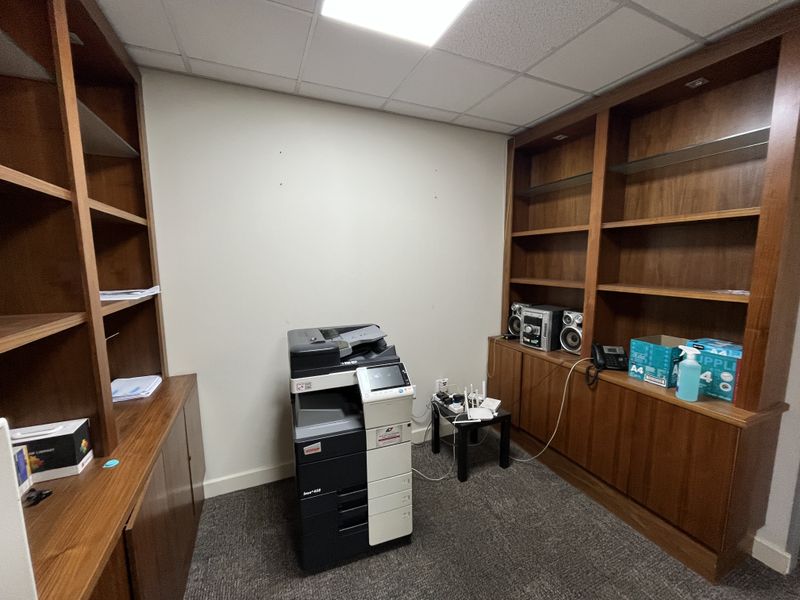 A functional office space featuring extensive built-in wooden shelving and cabinetry, carpeted flooring, and a suspended ceiling.