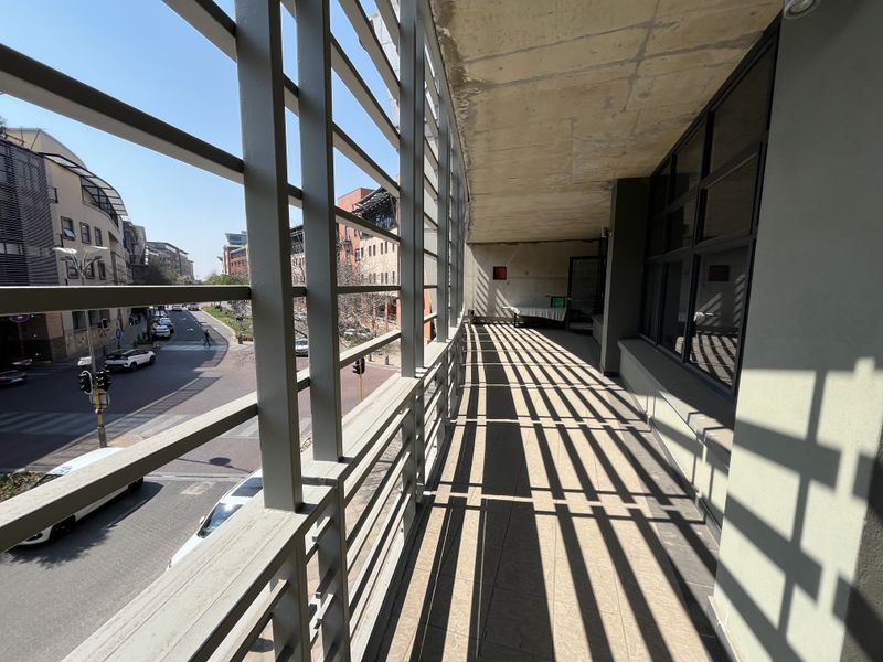 A spacious covered balcony featuring a metal balustrade, exposed concrete ceiling, and tiled flooring, offering an elevated view of the surrounding streetscape.