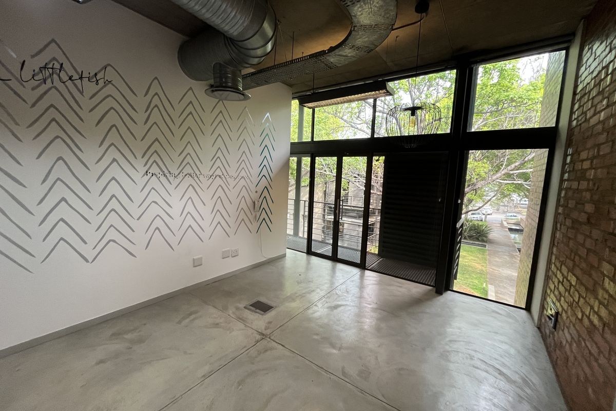 Modern open-plan office space featuring polished concrete floors, exposed industrial ducting, brick feature wall, and large windows providing natural light and outdoor views.