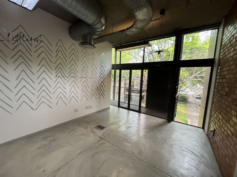 Modern open-plan office space featuring polished concrete floors, exposed industrial ducting, brick feature wall, and large windows providing natural light and outdoor views.