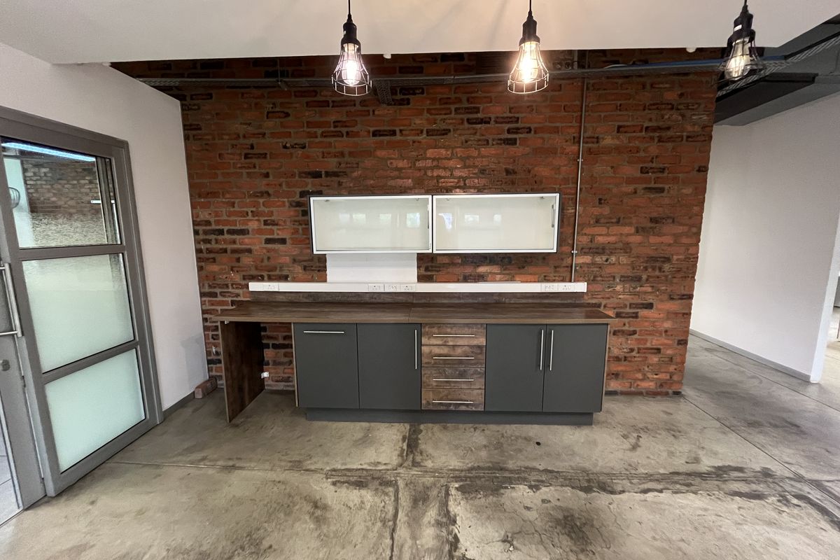 A modern office kitchenette featuring exposed brick walls, polished concrete flooring, dark cabinetry with wood accents, and industrial-style pendant lighting.