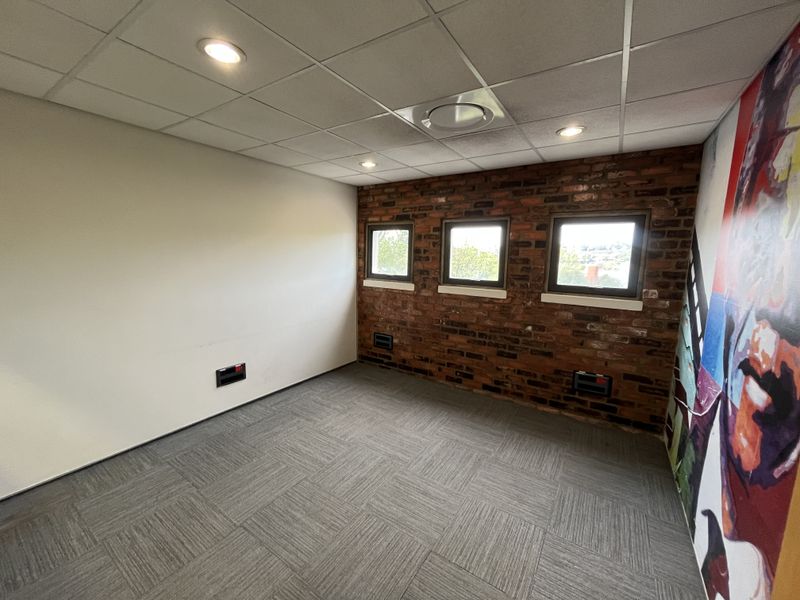 A bright, empty office space featuring an exposed brick feature wall, carpet tiles, suspended ceiling with recessed lighting, and multiple windows.