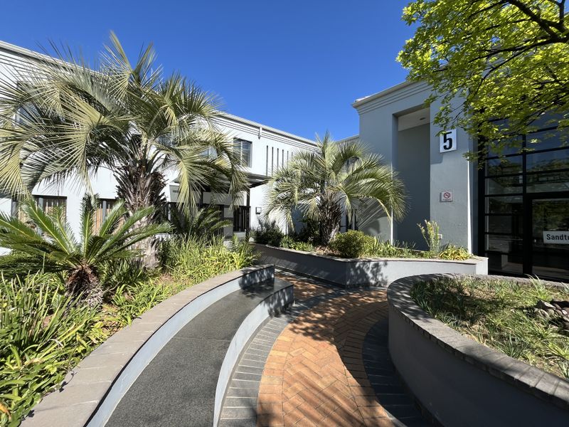 A well-maintained office building entrance featuring a curved brick paved walkway, landscaped garden beds, and modern glass facade elements.