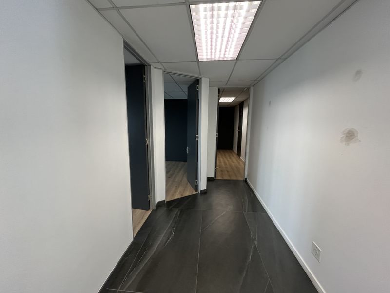 A functional office hallway featuring dark tiled flooring, suspended ceiling with recessed lighting, and access to multiple office rooms with laminate flooring.
