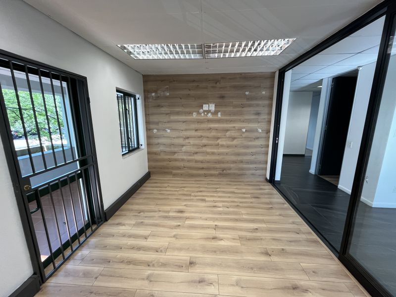 A bright, empty office space featuring wood-look flooring, a matching feature wall, recessed lighting, and a modern black-framed glass partition.