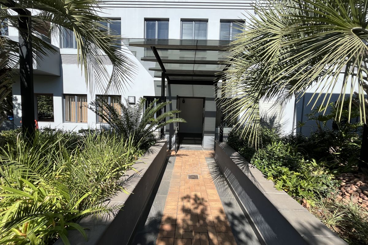 Front entrance of the office building featuring a glass canopy, brick paved walkway, and well-maintained landscaped garden beds.