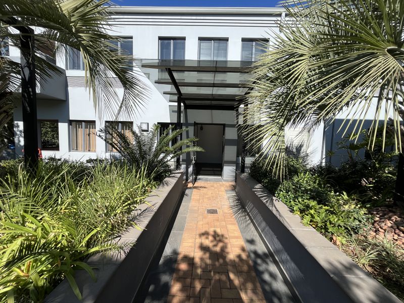 Front entrance of the office building featuring a glass canopy, brick paved walkway, and well-maintained landscaped garden beds.