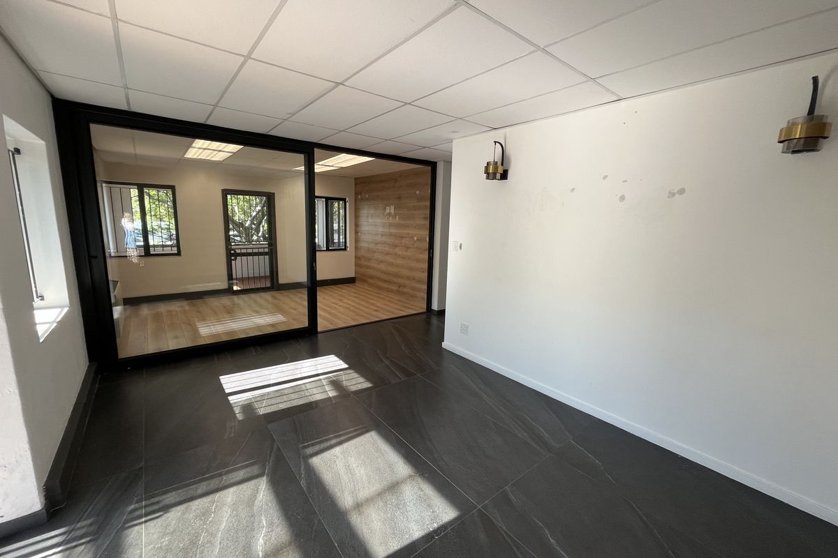 A bright, vacant office space featuring a modern glass partition, dark tiled flooring, and a suspended ceiling with wall-mounted lighting fixtures.