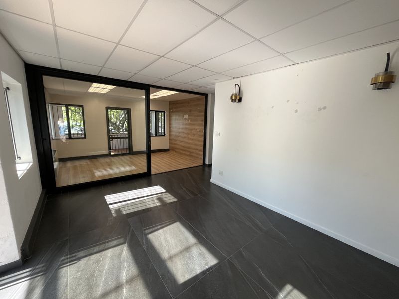 A bright, vacant office space featuring a modern glass partition, dark tiled flooring, and a suspended ceiling with wall-mounted lighting fixtures.