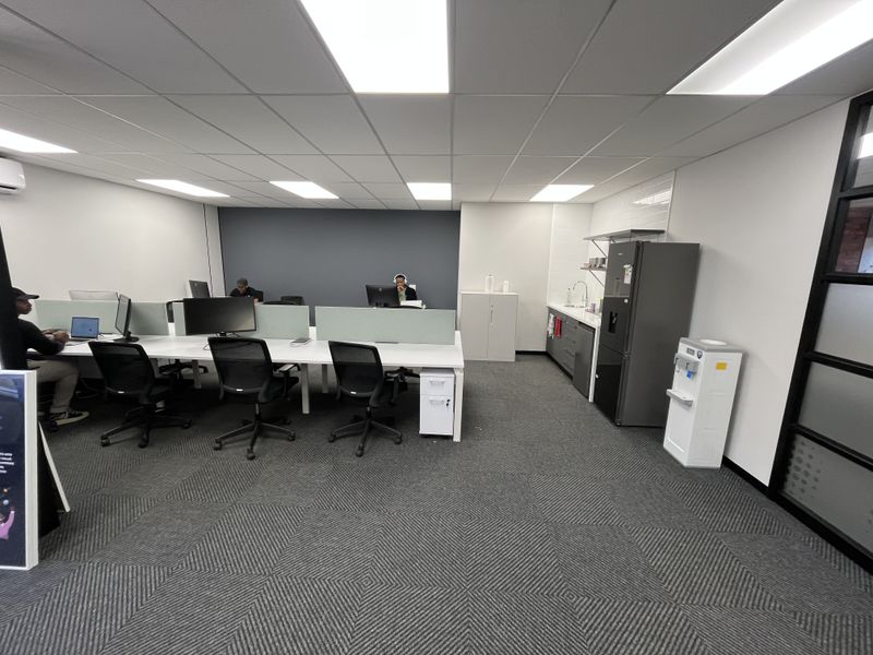 A modern, well-maintained office space featuring a partitioned workstation area, integrated kitchenette with sink, and professional suspended ceiling lighting.