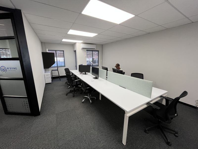 A modern, well-lit office space featuring a long partitioned desk setup, carpeted flooring, and an integrated air conditioning unit.