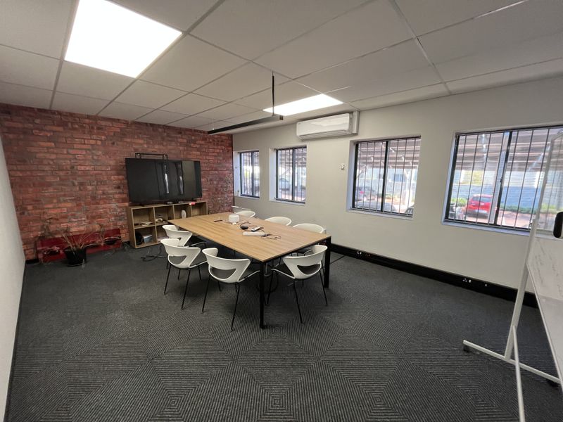 A functional office meeting room featuring an exposed brick wall, suspended ceiling with recessed lighting, split-system air conditioning, and security bars on the windows.