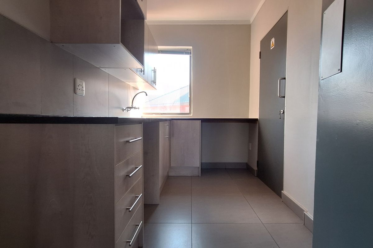 A clean, modern office kitchenette featuring light wood-finish cabinetry, dark countertops, tiled flooring, and a functional sink area.