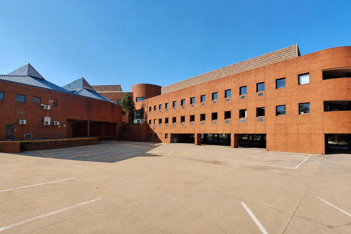 A large commercial office building featuring a distinctive red brick facade, multiple levels, and an expansive concrete parking area with covered bays.