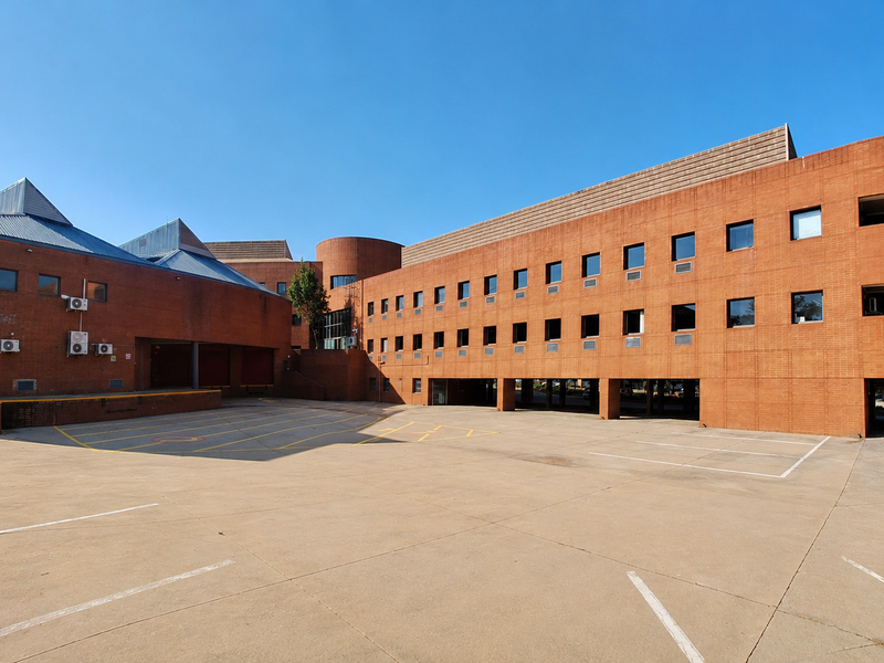 A large commercial office building featuring a distinctive red brick facade, multiple levels, and an expansive concrete parking area with covered bays.