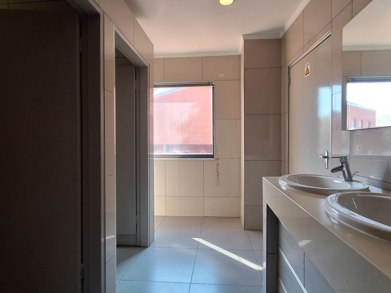A clean, modern office bathroom featuring dual vanity basins with chrome fixtures, neutral wall and floor tiling, and recessed lighting.