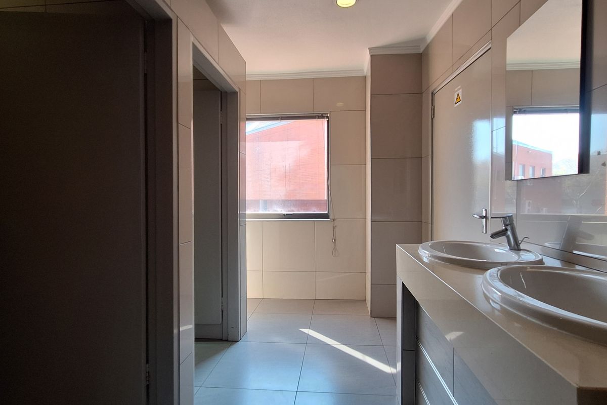 A clean, modern office bathroom featuring dual vanity basins with chrome fixtures, neutral wall and floor tiling, and recessed lighting.