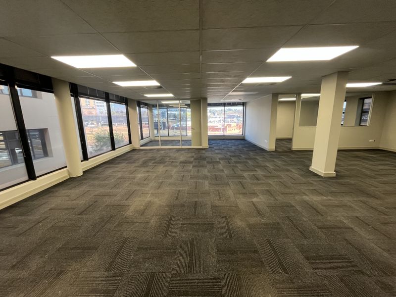 Spacious open-plan office area featuring large windows, carpet tiles, and a suspended ceiling with recessed lighting, offering a bright and professional workspace.