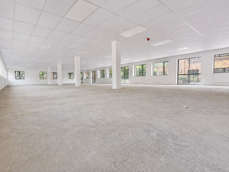 A spacious, open-plan office area featuring a suspended ceiling, structural columns, and large windows providing ample natural light.