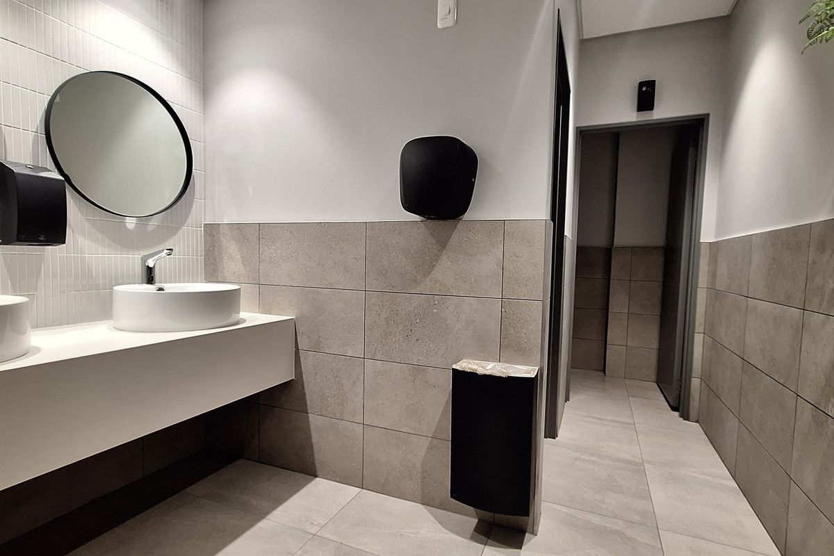 Modern office bathroom featuring dual vessel sinks, wall-mounted faucets, neutral tiled walls and flooring, and clean, contemporary finishes.