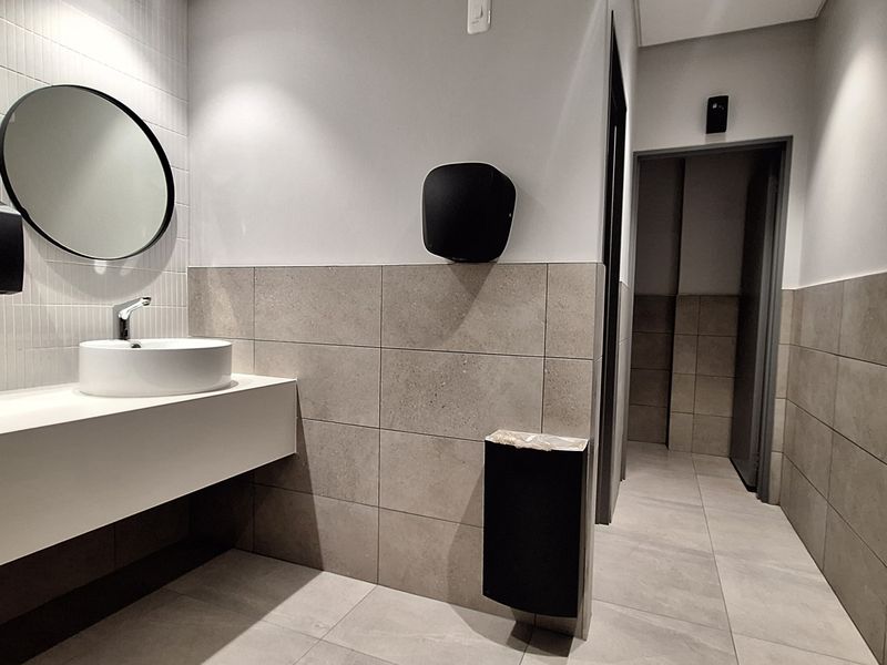 Modern office bathroom featuring dual vessel sinks, wall-mounted faucets, neutral tiled walls and flooring, and clean, contemporary finishes.