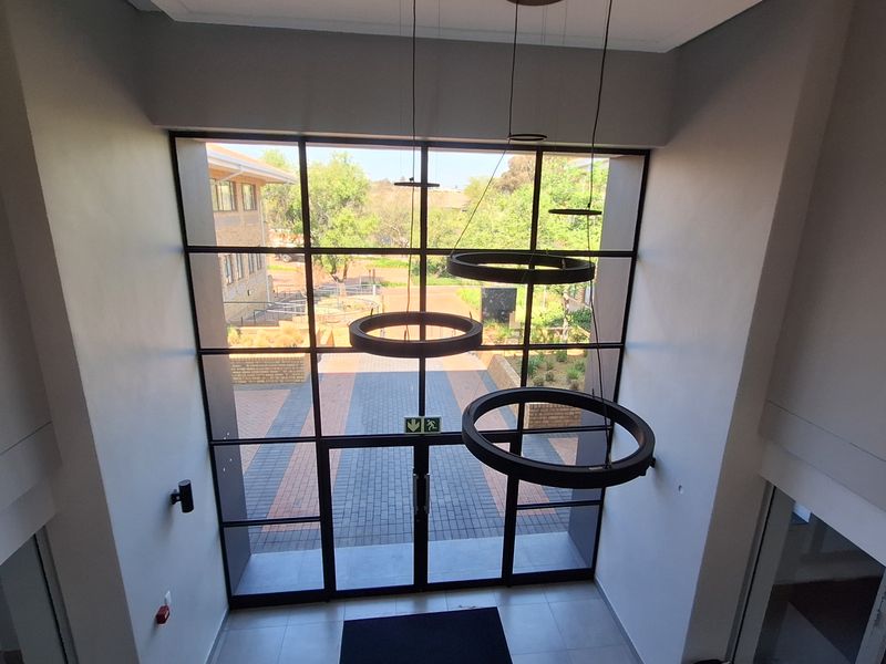 A bright, double-height office entrance featuring a large steel-framed glass window, modern ring pendant lighting, and clean tiled flooring.