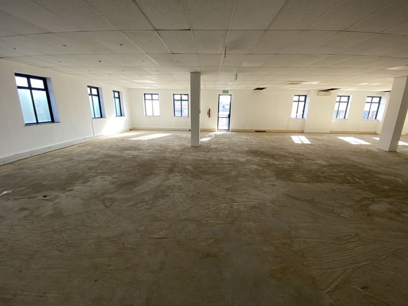 A large, open-plan office space featuring a suspended ceiling, multiple windows providing natural light, and unfinished concrete flooring ready for customization.