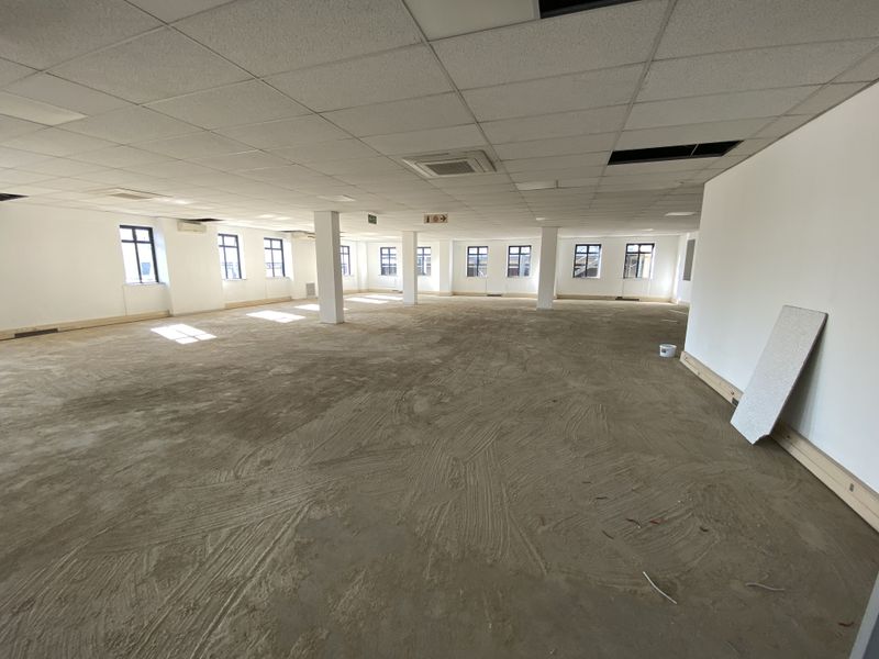 A large, open-plan office space featuring a suspended ceiling, structural columns, and large windows, currently undergoing renovation with unfinished flooring.