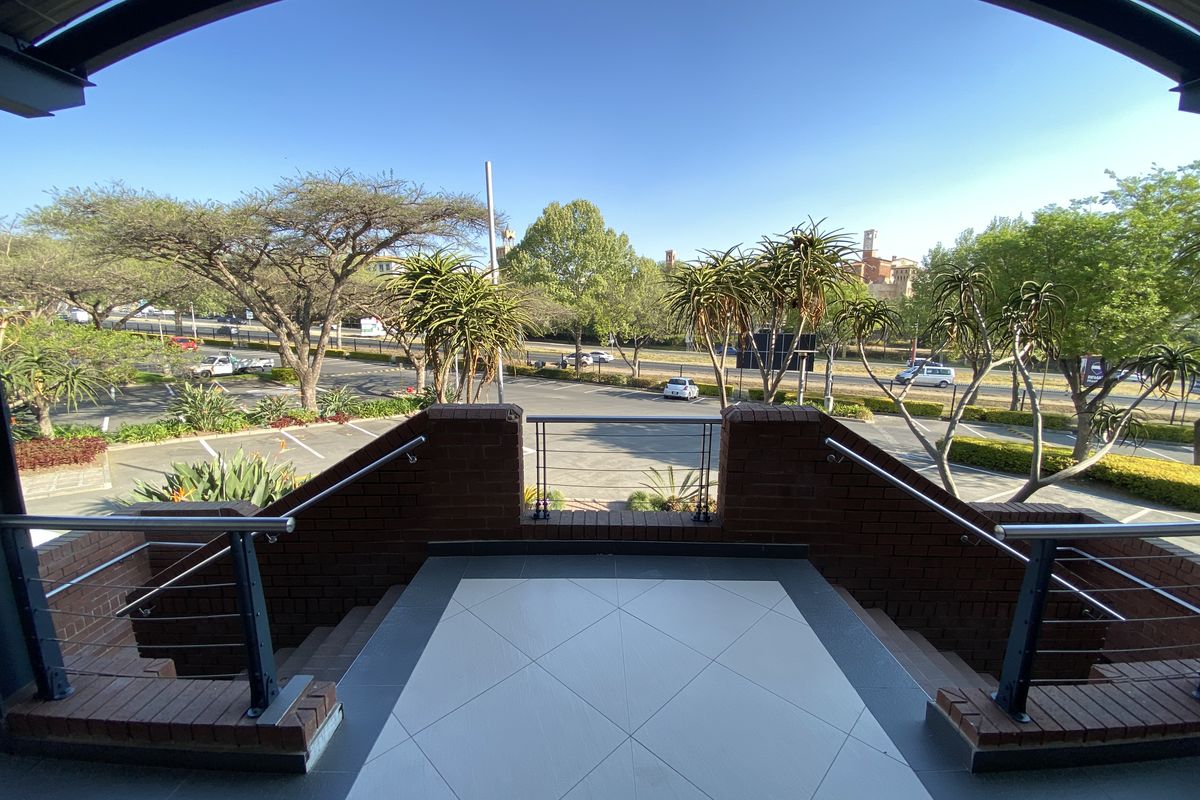 A covered balcony featuring brick balustrades, metal handrails, and tiled flooring, offering an elevated view of the surrounding office park and parking area.