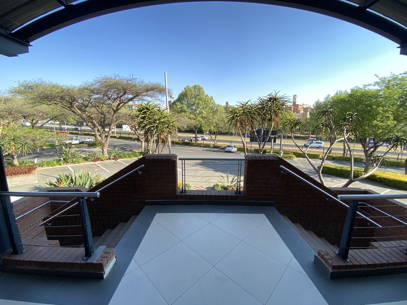 A covered balcony featuring brick balustrades, metal handrails, and tiled flooring, offering an elevated view of the surrounding office park and parking area.