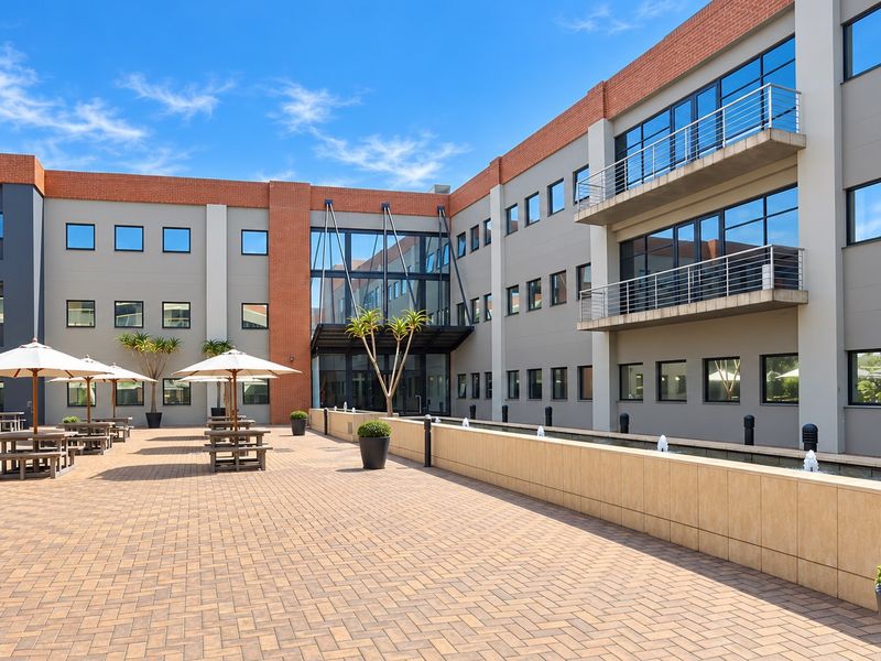 A modern office building exterior featuring a paved courtyard, a central water feature, and multi-level balconies with glass and brick architectural details.