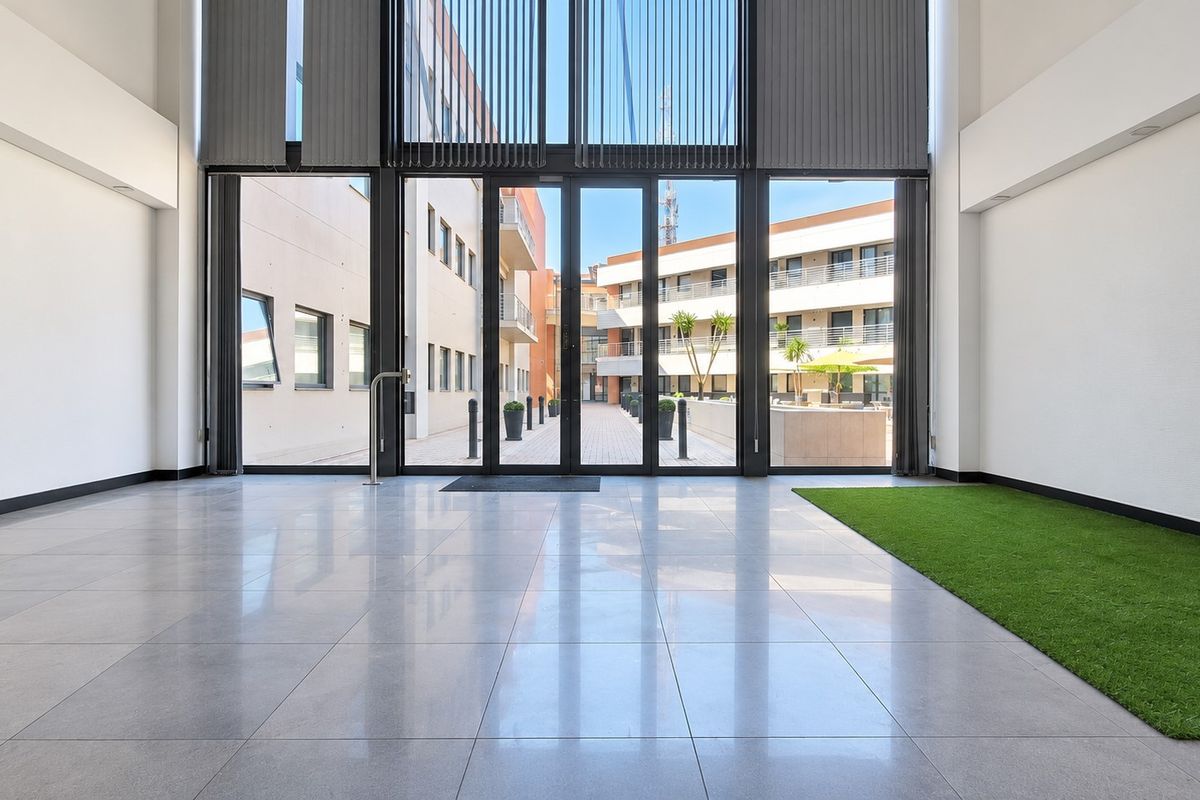 Spacious, modern office entrance hall featuring high ceilings, large glass sliding doors, and polished tiled flooring with an integrated artificial grass feature.
