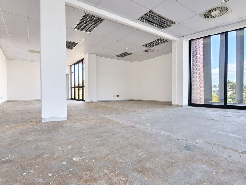 An open-plan office space featuring a suspended ceiling, recessed lighting, and large windows, currently awaiting flooring installation and final finishes.