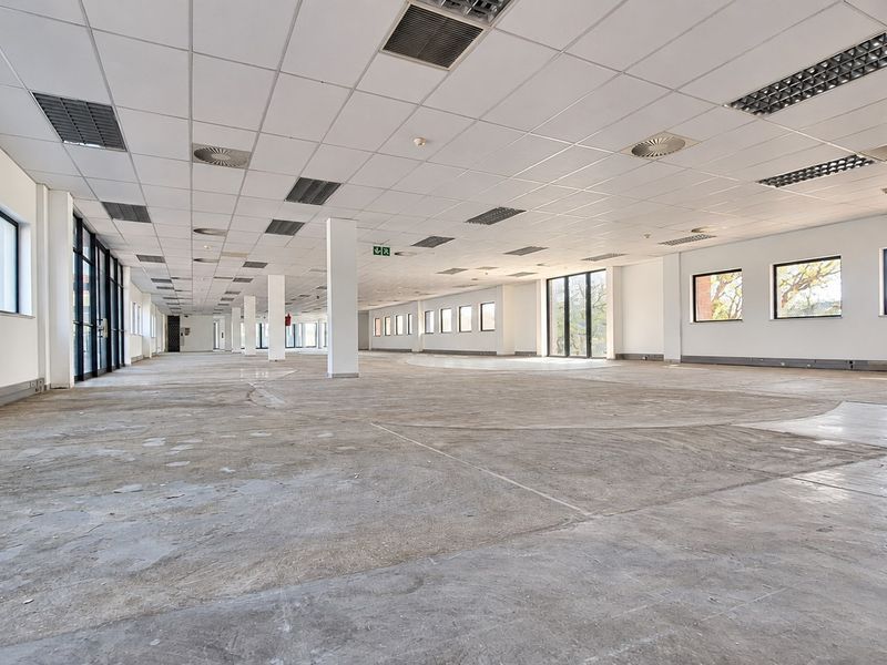 A large, open-plan office space featuring a suspended ceiling, recessed lighting, structural columns, and expansive windows, currently awaiting flooring installation.